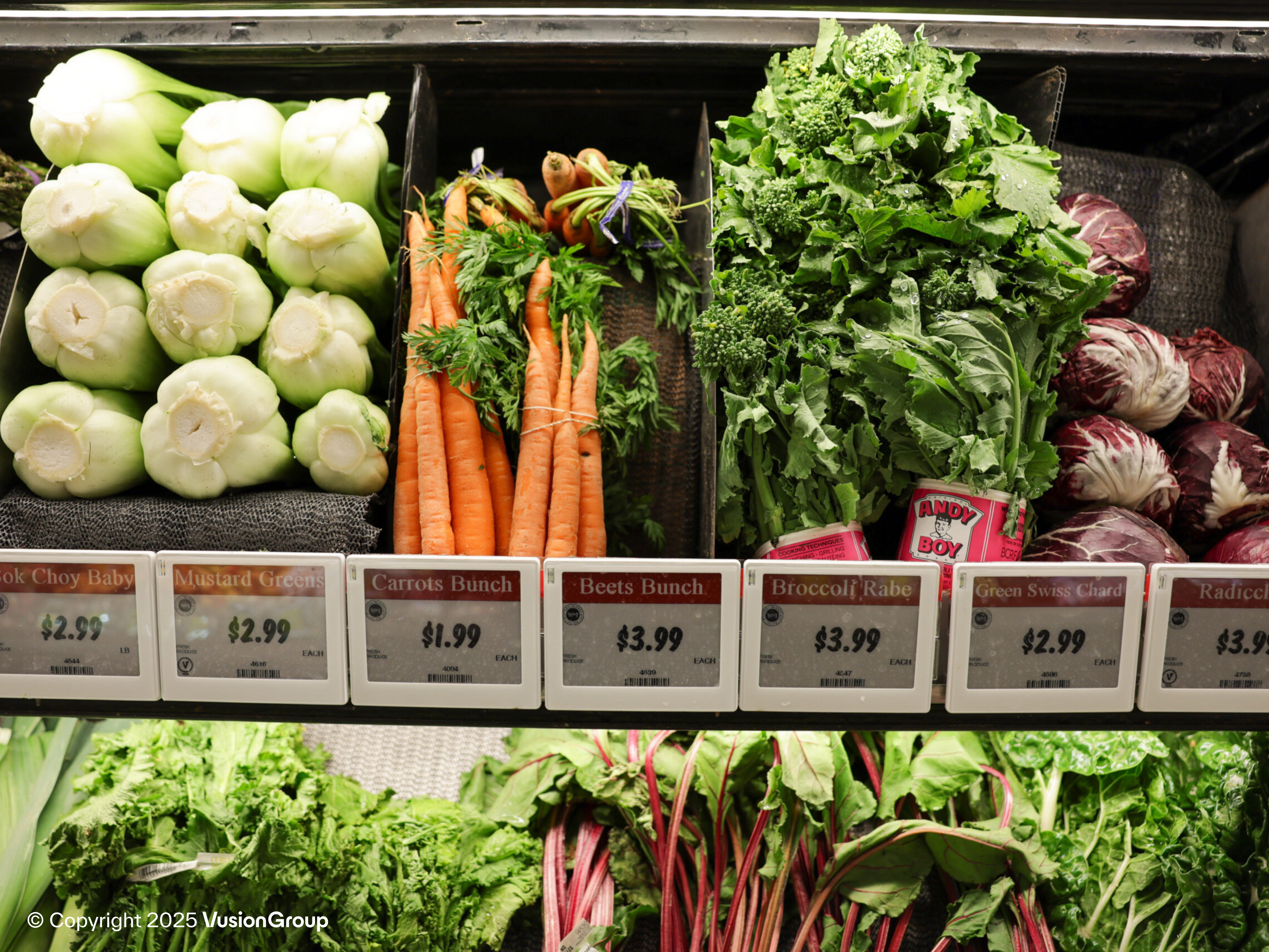 Close-up produce rail—carrots, bok choy, radicchio—showing synchronized electronic shelf labels integrated with DUMAC POS.